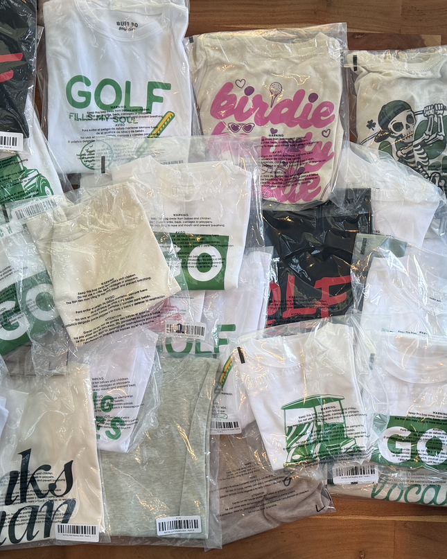 A pile of packaged t-shirts laid out on a wooden surface, many featuring golf-themed graphics and text such as 'GOLF', 'birdie', and 'GO'.