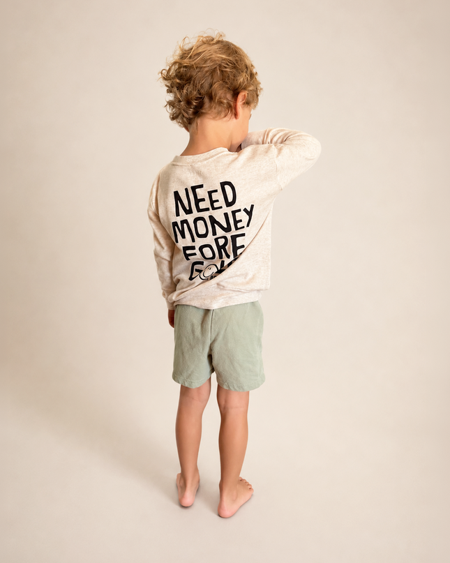 A child standing barefoot and facing away from the camera, wearing a light beige long-sleeve t-shirt with 'NEED MONEY FORE' printed in black on the back, paired with light green shorts, against a solid beige studio background.