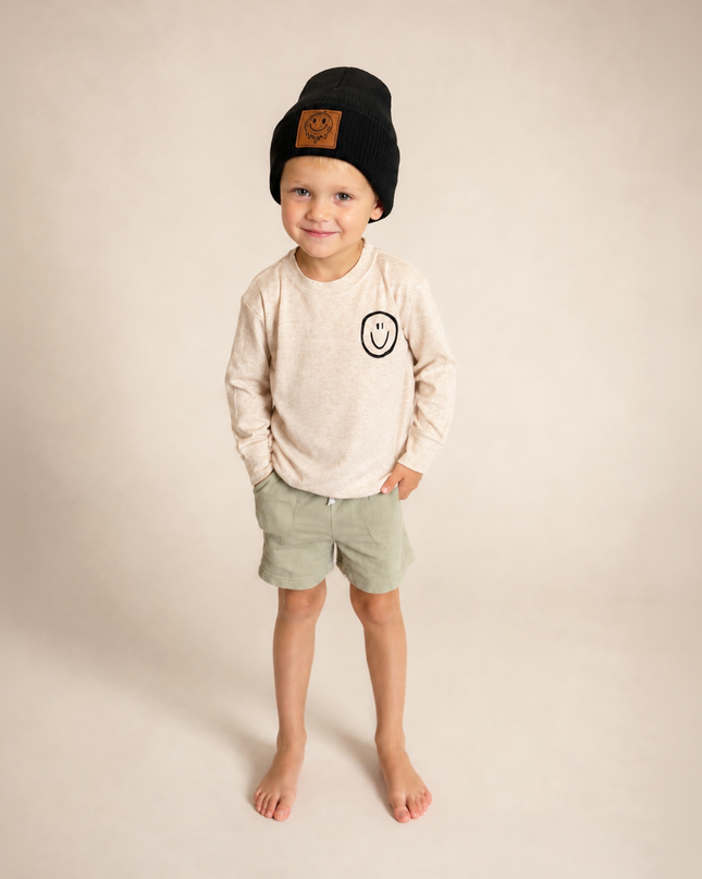 A young child standing barefoot against a plain beige studio background, wearing a black knit beanie with a brown patch, a light cream long-sleeve shirt with a small black smiley graphic on the chest, and light sage green shorts.