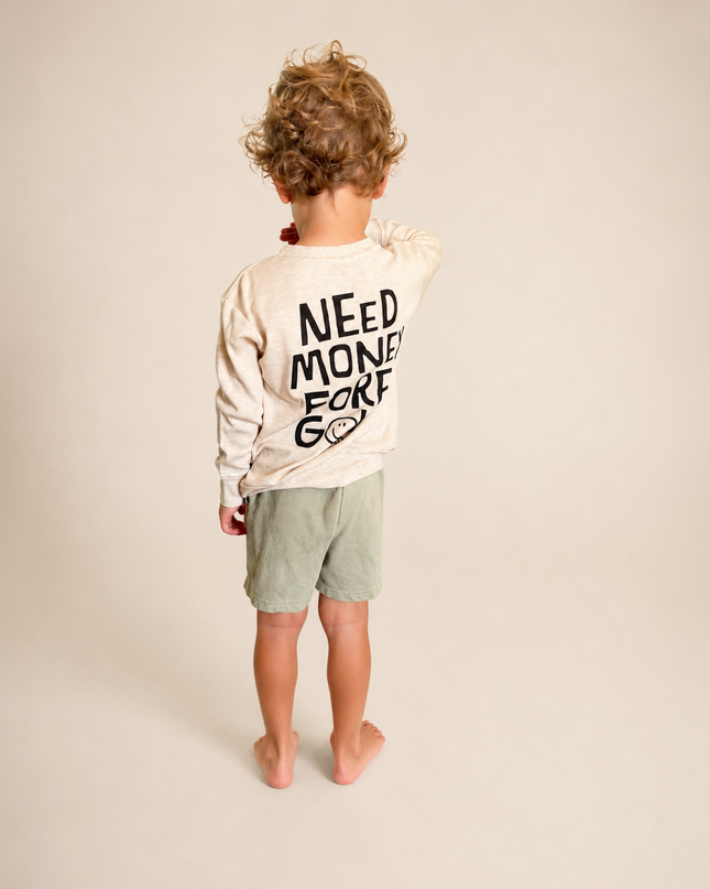 A toddler standing with their back to the camera, wearing a cream-colored long-sleeve shirt with black text reading 'NEED MONEY EAT GROW' and sage green shorts, against a solid light beige background.