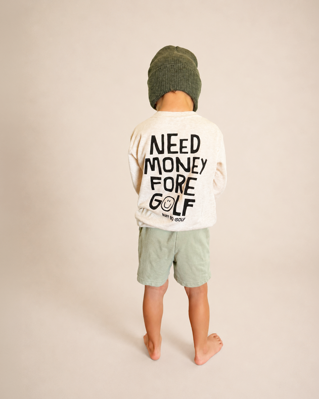 A child standing with their back to the camera, wearing a cream long-sleeve t-shirt with 'NEED MONEY FORE GOLF' printed in black on the back, sage green shorts, and a dark green knit beanie, against a solid beige studio background.