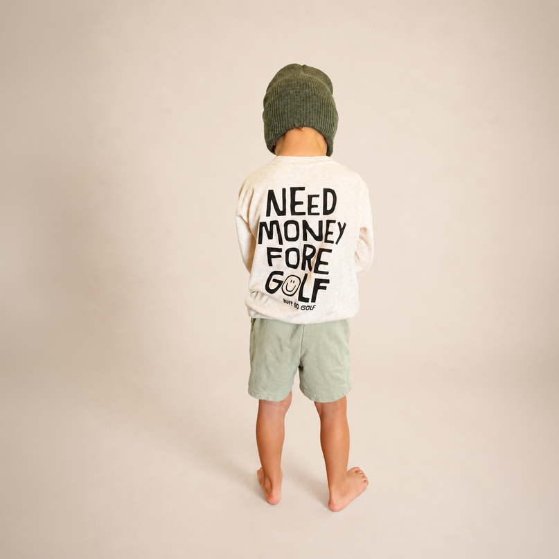 A child standing with their back to the camera, wearing a cream long-sleeve t-shirt with 'NEED MONEY FORE GOLF' printed in black on the back, sage green shorts, and a dark green knit beanie, against a solid beige studio background.