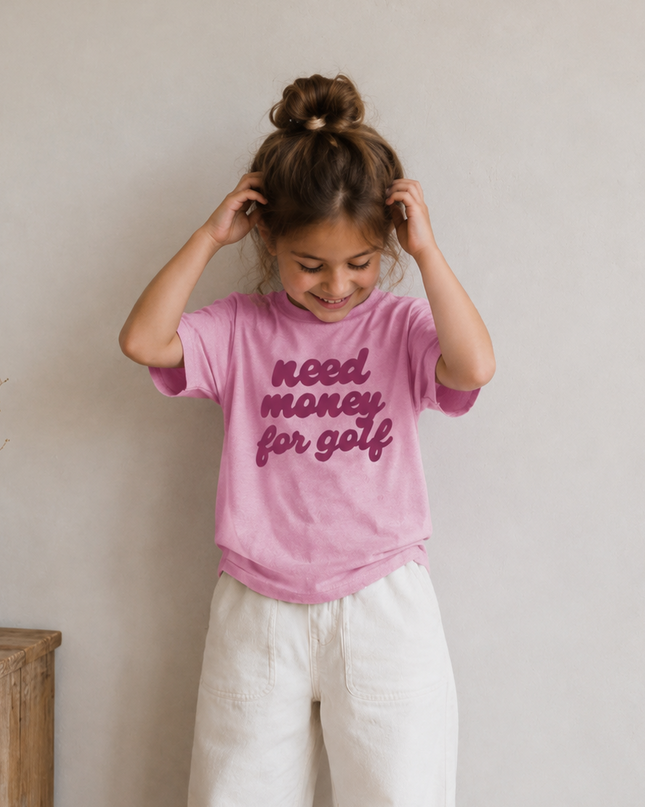 Child wearing a pink t-shirt with text, standing indoors against a plain wall.
