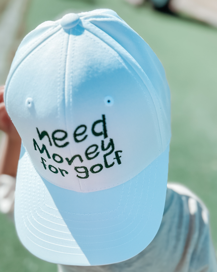 Child wearing a white baseball cap with playful embroidered text 'need money for golf' in dark green, standing on a golf course, fun youth golf accessory captured outdoors.