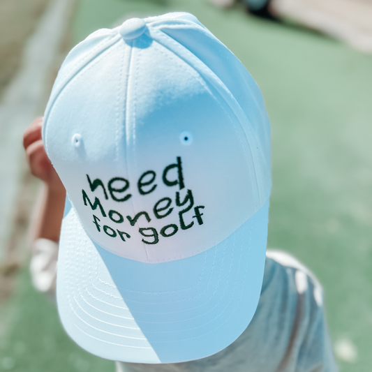 Child wearing a white baseball cap with playful embroidered text 'need money for golf' in dark green, standing on a golf course, fun youth golf accessory captured outdoors.