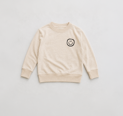 A light beige crewneck sweatshirt with a small black smiley face graphic on the left chest, laid flat against a light gray background.