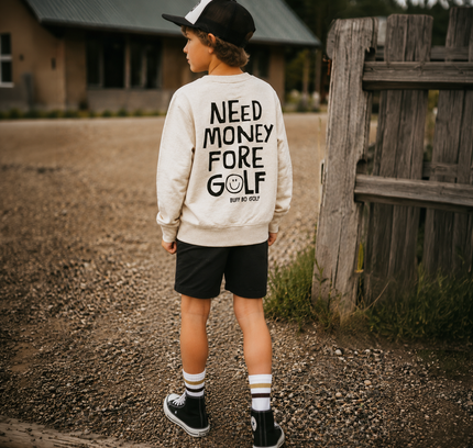 A child standing outdoors on a gravel path wearing a cream sweatshirt with bold black text reading 'NEED MONEY FORE GOLF' on the back, black shorts, striped socks, and black sneakers. A rustic wooden fence and a building are visible in the background.