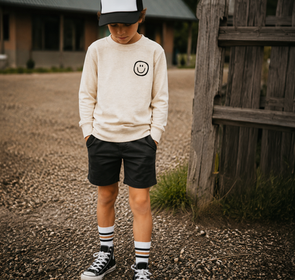 A child standing outdoors on a gravel path wearing a cream sweatshirt with a smiley face logo, black shorts, striped socks, and black-and-white sneakers, with a black-and-white trucker hat.