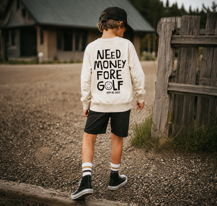 A child standing outdoors on a gravel path wearing a cream sweatshirt with bold black text reading 'NEED MONEY FORE GOLF', paired with black shorts, socks, and shoes, near a rustic wooden fence and cabin in the background.