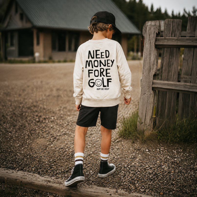 A child standing outdoors on a gravel path wearing a cream sweatshirt with bold black text reading 'NEED MONEY FORE GOLF', paired with black shorts, socks, and shoes, near a rustic wooden fence and cabin in the background.
