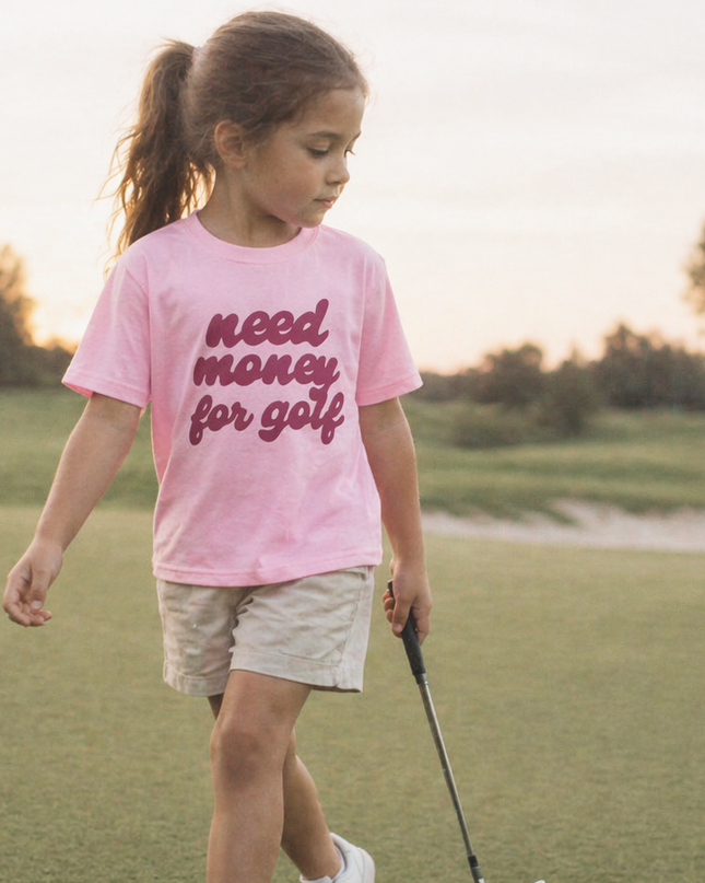 Young girl on a golf course wearing a pink t-shirt with text, holding a golf club.