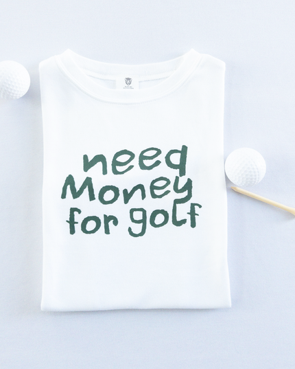 White kids golf t-shirt with playful dark green text reading 'Need Money for Golf,' displayed flat with golf balls and a wooden tee, fun and casual golf apparel for young golfers.