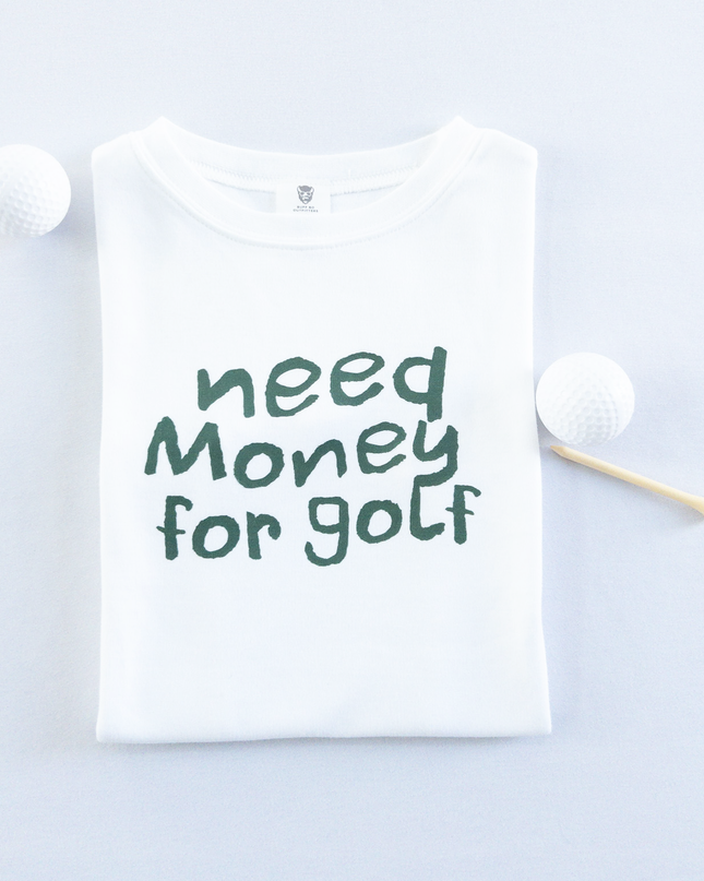 White kids golf t-shirt with playful dark green text reading 'Need Money for Golf,' displayed flat with golf balls and a wooden tee, fun and casual golf apparel for young golfers.