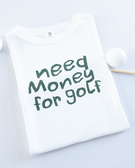 White kids golf t-shirt with playful dark green text reading 'Need Money for Golf,' displayed flat with golf balls and a wooden tee, fun and casual golf apparel for young golfers.