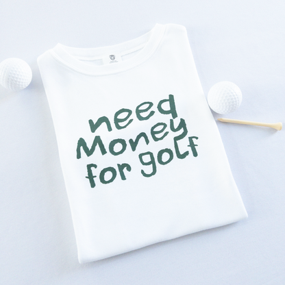 White kids golf t-shirt with playful dark green text reading 'Need Money for Golf,' displayed flat with golf balls and a wooden tee, fun and casual golf apparel for young golfers.