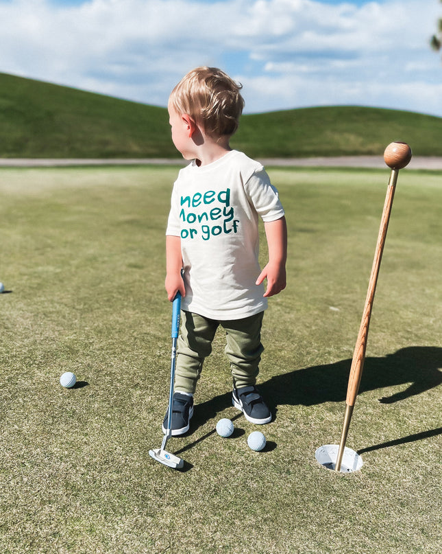 Young child putting on a golf green wearing a playful white t-shirt with dark green text reading 'Need Money for Golf,' surrounded by golf balls and a wooden flagstick, showcasing fun kids golf apparel outdoors.