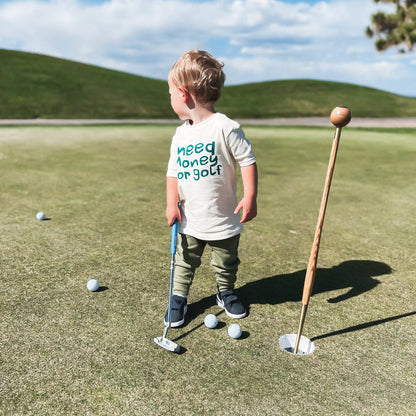 Young child putting on a golf green wearing a playful white t-shirt with dark green text reading 'Need Money for Golf,' surrounded by golf balls and a wooden flagstick, showcasing fun kids golf apparel outdoors.