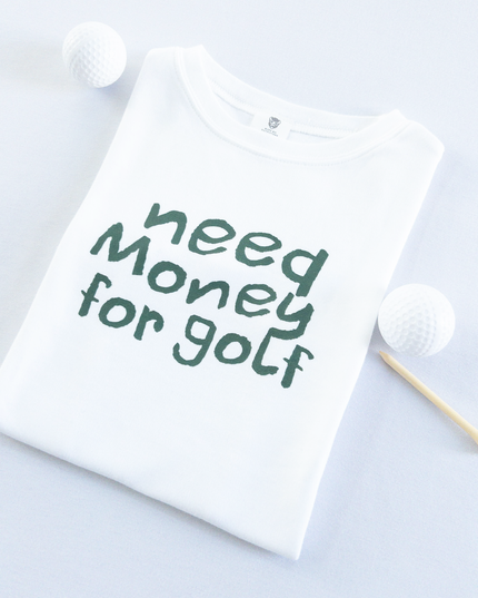 White kids golf t-shirt with playful dark green text reading 'Need Money for Golf,' displayed flat with golf balls and a wooden tee, fun and casual golf apparel for young golfers.