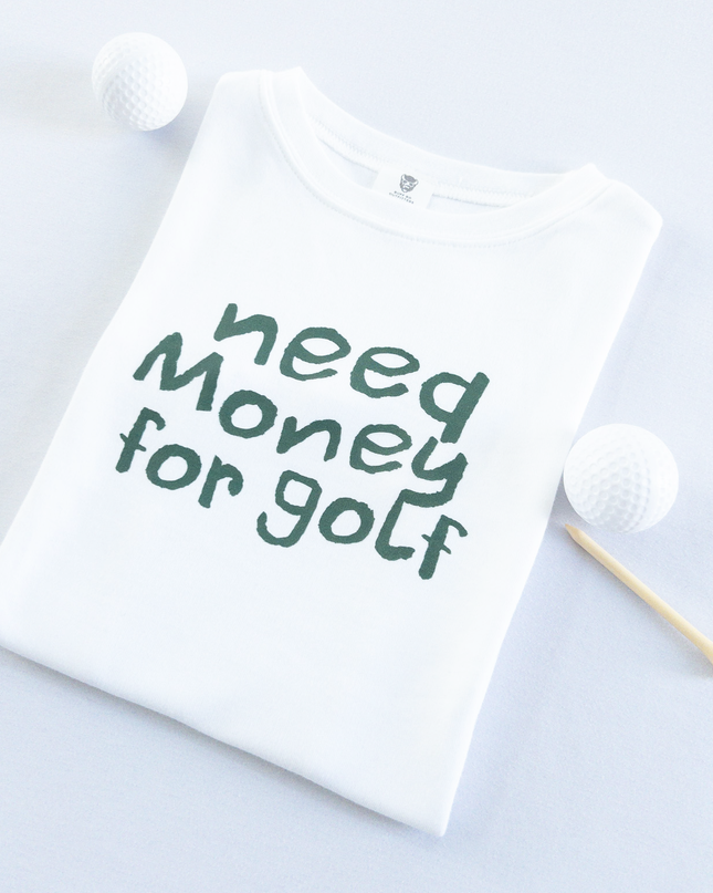 White kids golf t-shirt with playful dark green text reading 'Need Money for Golf,' displayed flat with golf balls and a wooden tee, fun and casual golf apparel for young golfers.