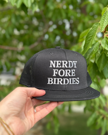 Black cap with 'NERDY FORE BIRDIES' text held by a hand against a blurred green background