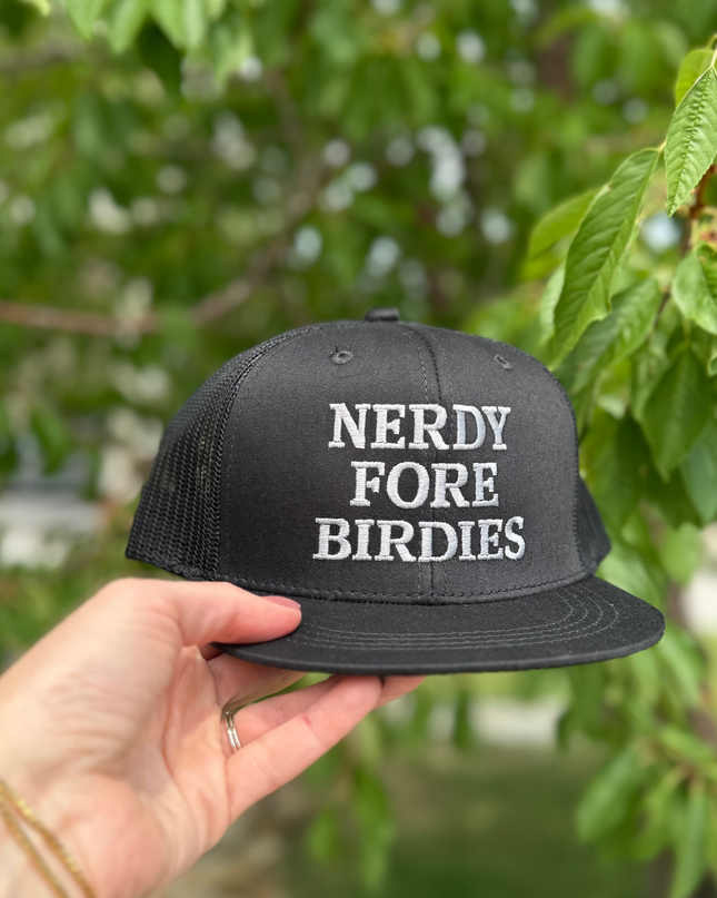Black cap with 'NERDY FORE BIRDIES' text held by a hand against a blurred green background