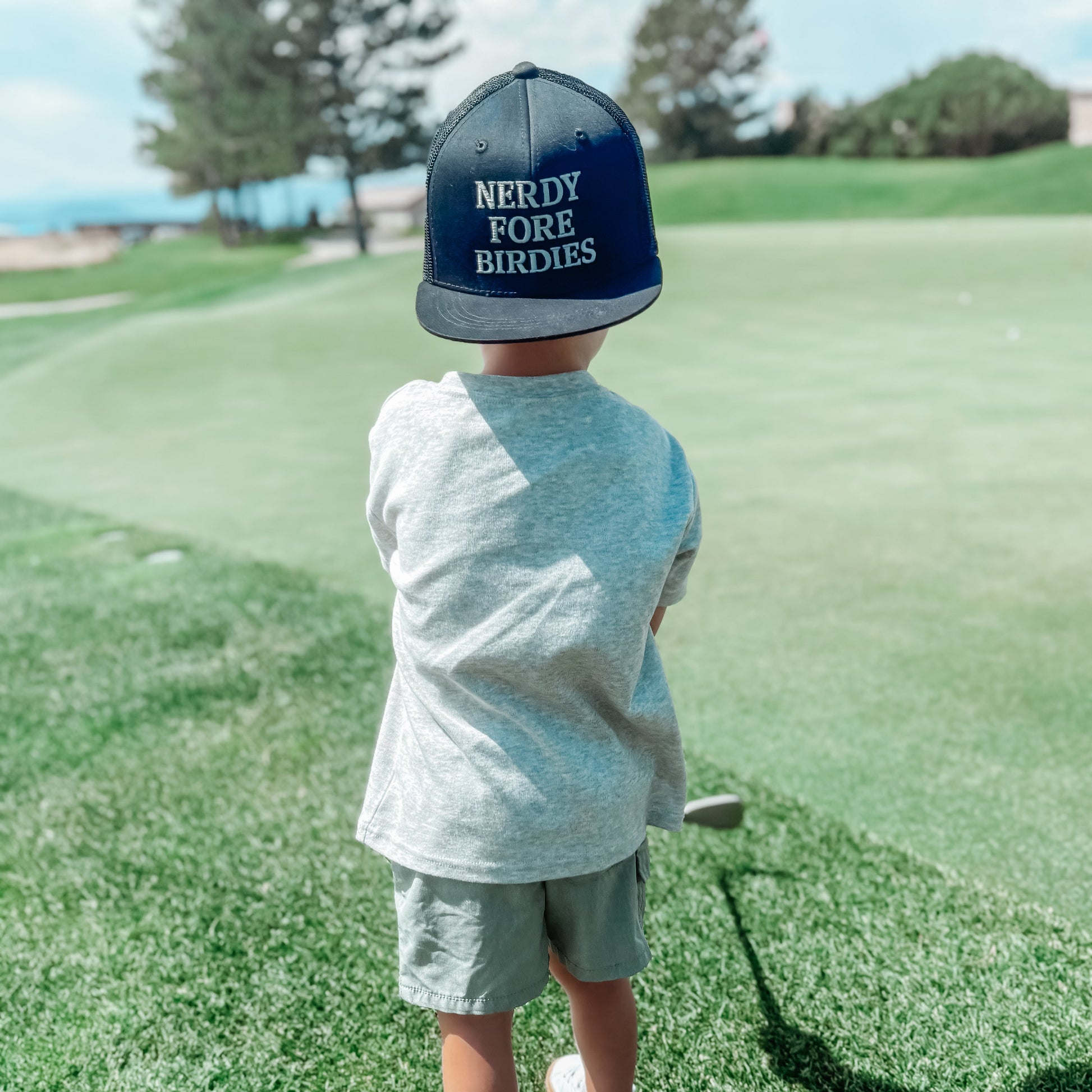 Child on a golf course wearing a cap with 'Nerdy Fore Birdies' text