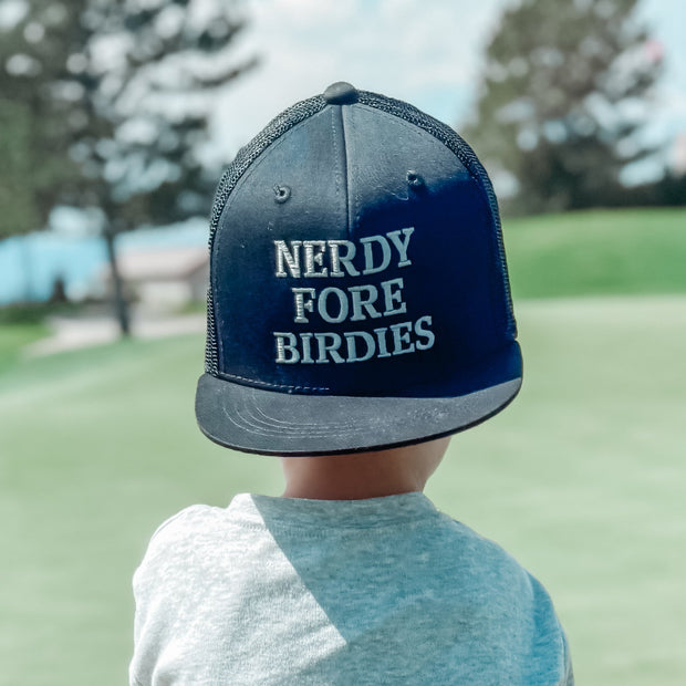 Child on a golf course wearing a cap with 'Nerdy Fore Birdies' text