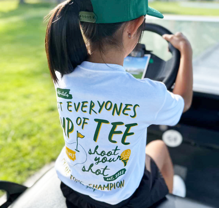 Person wearing a white t-shirt with green text and graphics, sitting in a car.