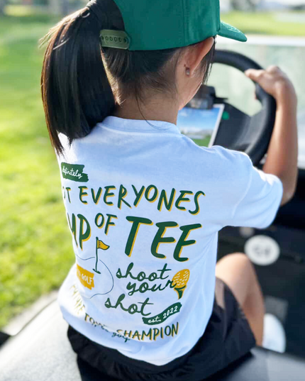 Person wearing a white t-shirt with green text and graphics, sitting in a car.