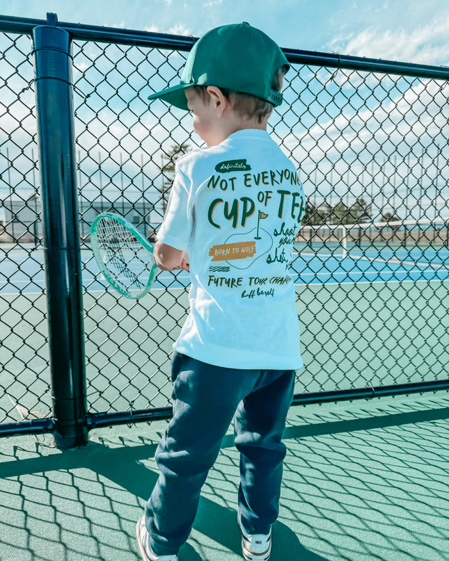 Child on a tennis court wearing a t-shirt with text, holding a tennis racket.