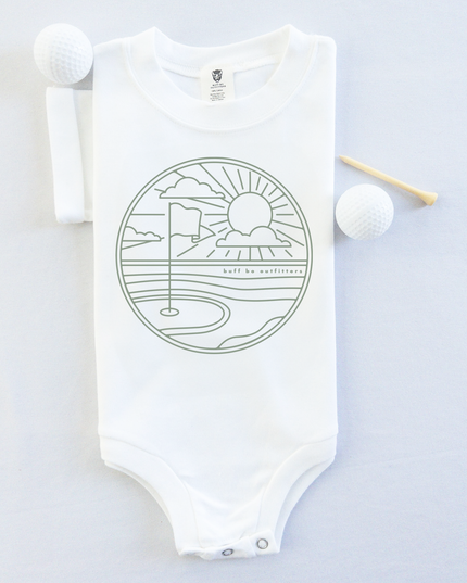 White short-sleeve baby onesie featuring a minimalist golf course design with a flag, sun, and clouds. The onesie is laid flat with two golf balls and a wooden tee beside it.