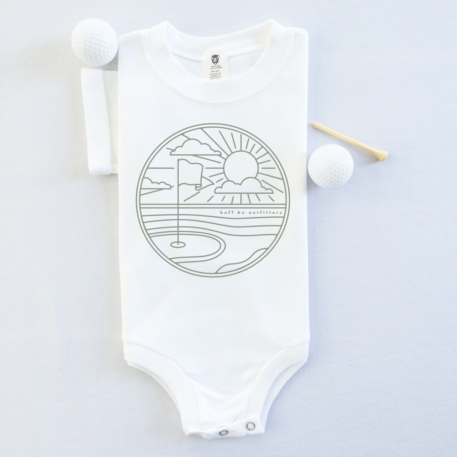 White short-sleeve baby onesie featuring a minimalist golf course design with a flag, sun, and clouds. The onesie is laid flat with two golf balls and a wooden tee beside it.