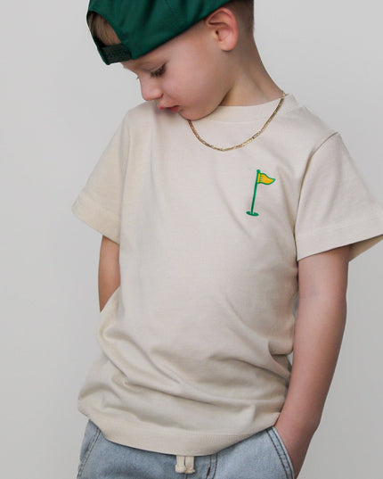 Child wearing a beige t-shirt with a small graphic and a green cap on a plain background