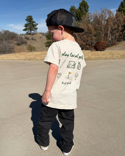 Child wearing a white t-shirt with graphics and text, standing on a paved surface with trees and clear sky in the background.