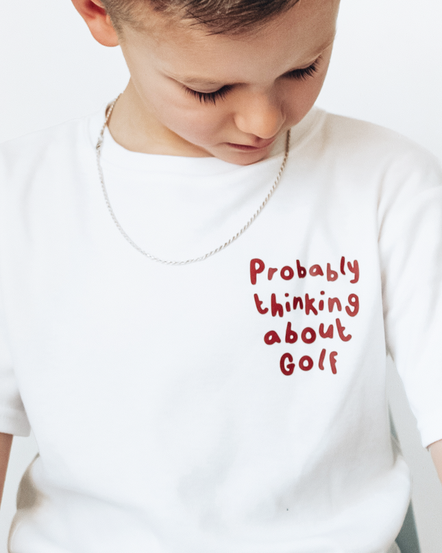 Child wearing a white t-shirt with red text 'Probably thinking about Golf' on a plain background