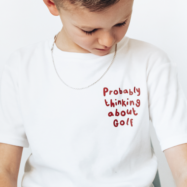 Child wearing a white t-shirt with red text 'Probably thinking about Golf' on a plain background