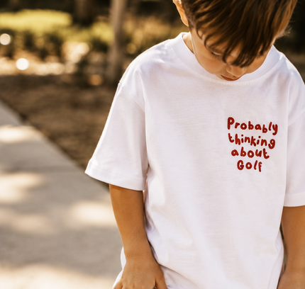 Child wearing a white t-shirt with red text 'Probably thinking about Golf' outdoors.
