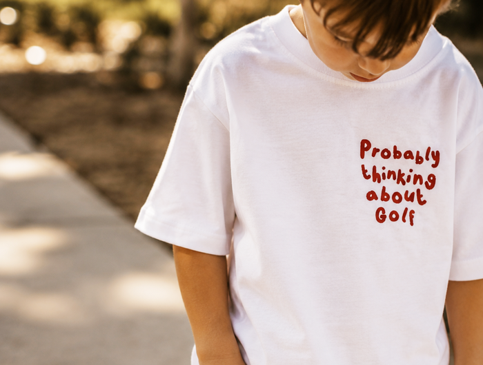 Child wearing a white t-shirt with red text 'Probably thinking about Golf' outdoors.