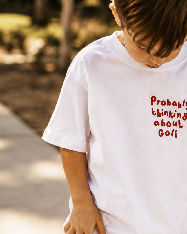 Child wearing a white t-shirt with red text 'Probably thinking about Golf' outdoors.