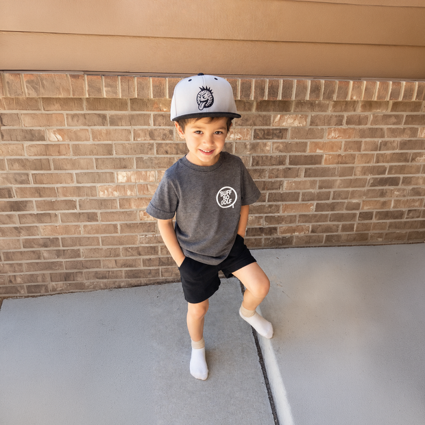 Child wearing a cap and t-shirt with a logo, standing against a brick wall.