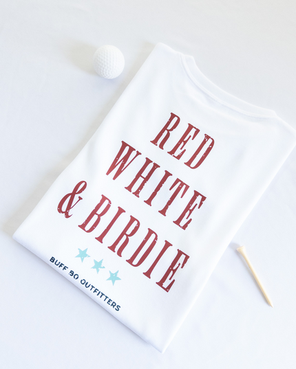 Folded white t-shirt with bold red text reading “RED WHITE & BIRDIE” and three light blue stars underneath, branded with “BUFF BO OUTFITTERS” at the bottom; styled with a white golf ball and wooden tee on a white background.
