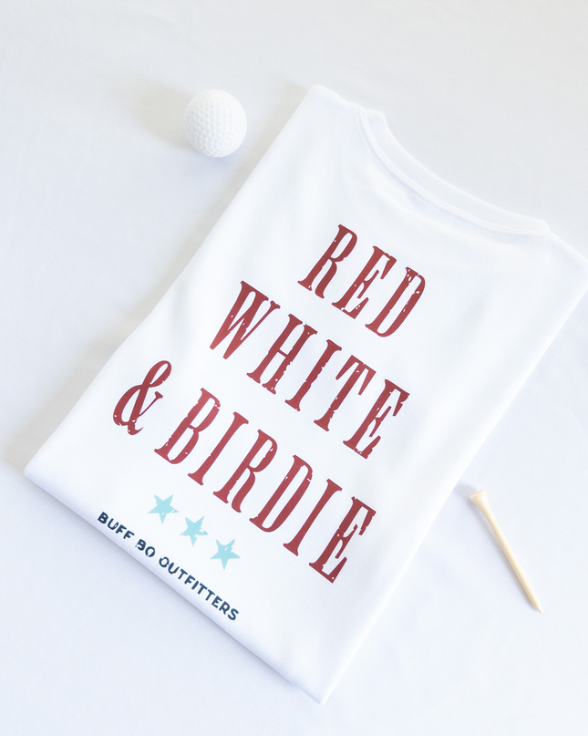 Folded white t-shirt with bold red text reading “RED WHITE & BIRDIE” and three light blue stars underneath, branded with “BUFF BO OUTFITTERS” at the bottom; styled with a white golf ball and wooden tee on a white background.
