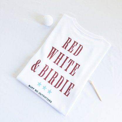 Folded white t-shirt with bold red text reading “RED WHITE & BIRDIE” and three light blue stars underneath, branded with “BUFF BO OUTFITTERS” at the bottom; styled with a white golf ball and wooden tee on a white background.
