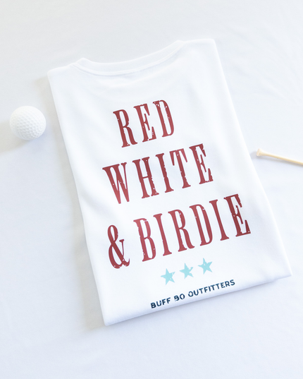 Folded white t-shirt with bold red text reading “RED WHITE & BIRDIE” and three light blue stars underneath, branded with “BUFF BO OUTFITTERS” at the bottom; styled with a white golf ball and wooden tee on a white background.
