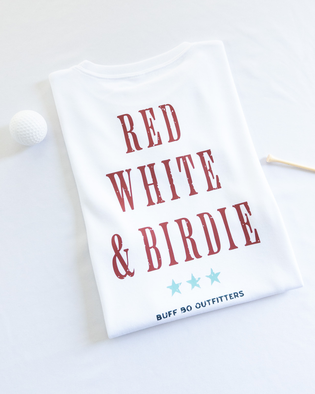 Folded white t-shirt with bold red text reading “RED WHITE & BIRDIE” and three light blue stars underneath, branded with “BUFF BO OUTFITTERS” at the bottom; styled with a white golf ball and wooden tee on a white background.
