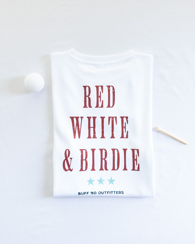 Folded white t-shirt with bold red text reading “RED WHITE & BIRDIE” and three light blue stars underneath, branded with “BUFF BO OUTFITTERS” at the bottom; styled with a white golf ball and wooden tee on a white background.
