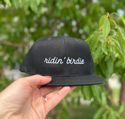 Black cap with 'ridin' birdie' text held by a hand against a green leafy background