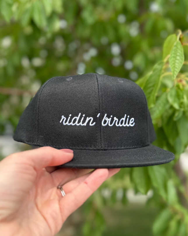 Black cap with 'ridin' birdie' text held by a hand against a green leafy background