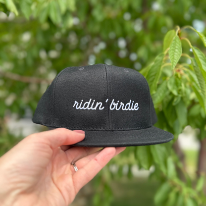 Black cap with 'ridin' birdie' text held by a hand against a green leafy background