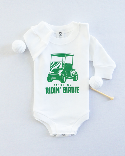 White baby golf onesie featuring a green golf cart graphic and playful text reading 'Catch Me Ridin' Birdie,' styled flat with two golf balls and a wooden tee, fun golf-themed outfit for babies and toddlers.